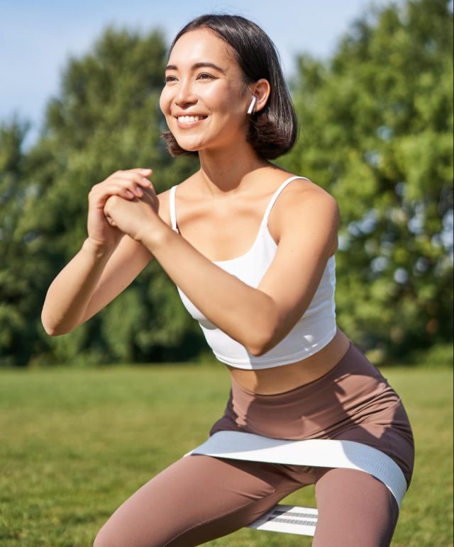 Asian fitness girl doing squats in park using resistance band.